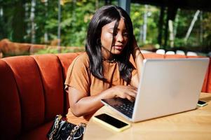 ritratto di bellezza giovane nero donna, indossare arancia vestito, seduta con il computer portatile su ufficio. foto