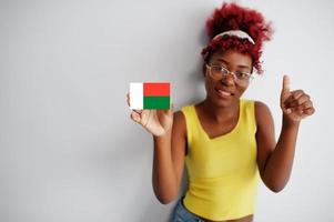 africano donna con afro capelli, indossare giallo canottiera e occhiali, hold Madagascar bandiera isolato su bianca sfondo, mostrare pollice su. foto