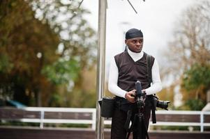 giovane professionale africano americano operatore video Tenere professionale telecamera con professionista attrezzatura. afro cineoperatore indossare nero duraq fabbricazione un' video. foto