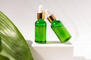 verde bicchiere cosmetico bottiglie con un' contagocce su un' bianca podio con tropicale le foglie. naturale cosmetici concetto, naturale essenziale olio e pelle cura prodotti foto