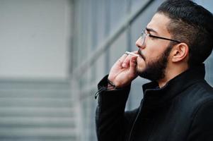mezzo orientale imprenditore indossare nero cappotto e blu camicia, occhiali contro ufficio edificio fumo sigaretta. foto