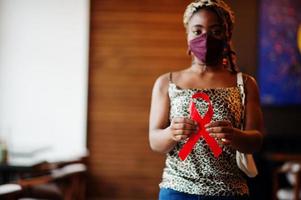 AIDS consapevolezza. elegante africano americano donna con dreadlocks afro capelli, indossare viso proteggere maschera e copricapo leopardo sciarpa, hold rosso nastro. foto