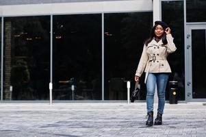 alla moda africano americano donna indossare berretto e cappotto con borsetta in posa all'aperto. foto