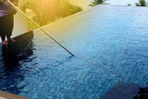 donna lavoratore pulito privato nuoto piscina con vuoto blu tubo addetto alle pulizie ogni settimana nel estate, sale genere piscina lungo con oceano come orizzontale stile con Noce di cocco alberi foto