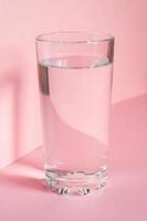 vicino su di scintillante acqua nel trasparente alto bicchiere e sole bagliore su rosa sfondo foto