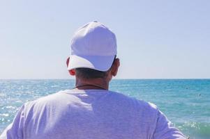 medio anziano uomo guardare per il oceano. indietro di il uomo nel di Pentecoste berretto e maglietta foto