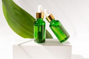 verde bicchiere cosmetico bottiglie con un' contagocce su un' bianca podio con tropicale le foglie. naturale cosmetici concetto, naturale essenziale olio e pelle cura prodotti foto