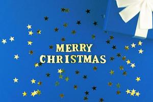 di legno lettere su blu sfondo. allegro Natale lettering su blu carta con sparpagliato stelle coriandoli e regalo scatola. foto