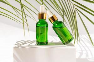 verde bicchiere cosmetico bottiglie con un' contagocce su un' bianca podio con tropicale le foglie. naturale cosmetici concetto, naturale essenziale olio e pelle cura prodotti foto