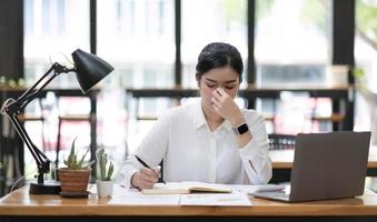 giovane donna asiatica che sente ceppo di testa di emicrania. finanziera donna d'affari stanca e oberata di lavoro mentre lavorava su un computer portatile in ufficio. foto