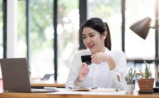 giovane asiatico donna d'affari avere il piacere di Guardando opera su loro il computer portatile e preferito caffè a il ufficio. foto