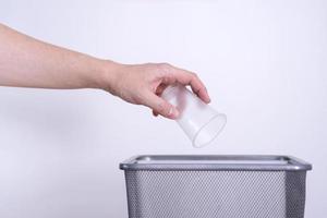 un' mano lanci un' plastica monouso tazza in il spazzatura contro un' grigio sfondo. foto