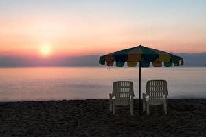 sedia a sdraio bianca e ombrellone sulla spiaggia tropicale con cielo arancione foto