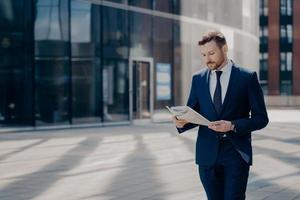 concentrato uomo d'affari vestito formalmente che legge il giornale mentre cammina vicino all'edificio per uffici foto