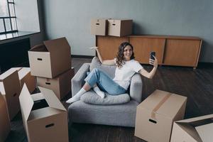 felice ragazza spagnola seduta in poltrona e fare una videochiamata sullo smartphone. proprietario in appartamento nuovo. foto