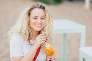 bella giovane donna sorridente con capelli ricci chiari, sorriso a trentadue denti, ricrea sulla spiaggia, beve un cocktail fresco estivo, vestita con una maglietta bianca casual, riposa bene. persone, estate, concetto di stile di vita foto