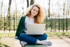 donna caucasica con capelli ricci chiari e occhi azzurri che indossa abiti comodi seduta a gambe incrociate all'aperto tenendo il laptop in ginocchio messaggiando con il suo ragazzo guardando direttamente nella fotocamera foto