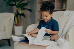 felice ragazzino di razza mista con graziosi capelli ricci che legge un libro a casa foto
