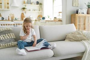 poco ragazzo ragazza bambino in età prescolare leggere fiaba libro seduta su accogliente divano a casa solo. bambino formazione scolastica foto