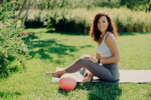 donna bruna positiva in top corto e leggings posa a piedi nudi su karemat usa la palla per esercizi ginnici sorride piacevolmente gode di una splendida vista sulla natura. sport all'aperto. pratica yoga. foto