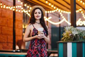 carina donna bruna che indossa cappello e vestito, in piedi alla caffetteria, con in mano una tazza di carta con caffè o tè. giovane imprenditrice a cena al bar che riposa dopo un duro lavoro in ufficio. concetto di bellezza foto