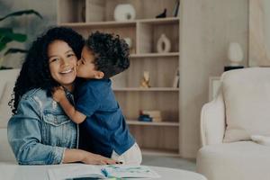 Ritratto di ragazzo carino afroamericano in maglietta polo blu che abbraccia la madre e la bacia sulla guancia foto