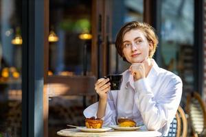 donna caucasica sorseggiando un caffè espresso caldo mentre si siede fuori dal bistrot del caffè in stile europeo godendosi la vita lenta con l'atmosfera mattutina nella piazza della città con pasticceria dolce foto