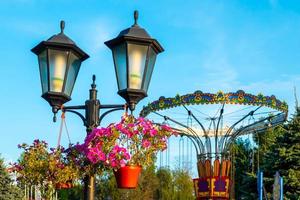 paesaggio di un divertimento parco con lanterna decorato con fiori sfondo il superiore di un' Ferris ruota mostrando sopra il albero cime contro un' cielo. foto