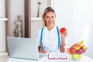 ritratto di giovane sorridente femmina dietologo nel il consultazione camera. dietologo scrivania con salutare frutta, succo e misurazione nastro. dietista Lavorando su dieta Piano. foto