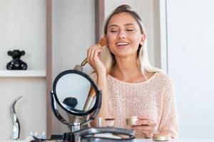 bellezza donna l'applicazione trucco. bellissimo ragazza guardare nel il specchio e l'applicazione cosmetico con un' grande spazzola. ragazza prende arrossire su il zigomi. polvere, rossetto foto