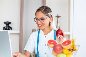 medico dietologo scrittura Astuccio storia nel il ufficio. giovane donna dietista prescrivere ricetta. femmina dietologo seduta a tavolo con il computer portatile foto