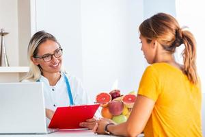 medico nutrizionista, dietista e femmina paziente su consultazione nel il ufficio. giovane sorridente femmina dietologo nel il consultazione camera. dietologo scrivania con salutare frutta e misurazione nastro. foto