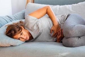 donna nel doloroso espressione Tenere mani contro pancia sofferenza mestruale periodo dolore, dire bugie triste su casa letto, avendo pancia crampo nel femmina Salute concetto foto