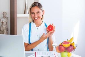 ritratto di giovane sorridente femmina dietologo nel il consultazione camera. dietologo scrivania con salutare frutta, succo e misurazione nastro. dietista Lavorando su dieta Piano. foto