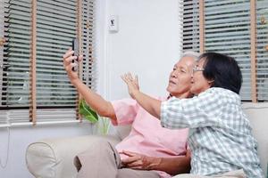 asiatico anziano coppia incontrare nel virtuale famiglia, bambini e nipoti attraverso mobile telefoni durante coronavirus. sociale distanza. contento sorridente coppia condivisione loro smartphone su il divano a casa. foto