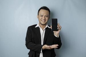 un' ritratto di un' contento asiatico uomo d'affari è sorridente e Tenere il suo smartphone mostrando copia spazio su è schermo indossare nero completo da uomo isolato di un' blu sfondo foto