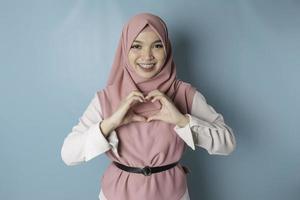 un attraente giovane asiatico musulmano donna indossare un' rosa hijab si sente contento e un' romantico forme cuore gesto esprime tenero sentimenti foto