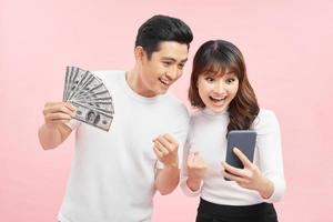 attraente giovane coppia in piedi insieme isolato al di sopra di rosa sfondo, mostrando i soldi banconote, utilizzando mobile Telefono foto