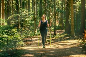 donna escursionismo. passeggiate attraverso il denso foresta utilizzando il trekking poli. Sorridi su il suo viso e occhiali da sole. di sole raggi rompere attraverso il foresta foto