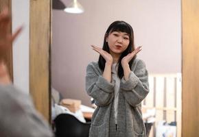 carino asiatico coreano ragazza sceglie Abiti, prende cura di se stessa e pose divertente nel davanti di il specchio nel il vivente camera. foto