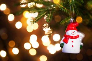 Natale giocattolo pupazzo di neve nel un' rosso cappello con un' sciarpa su vivere ramo di un' abete albero con d'oro luci di ghirlande nel sfocatura. nuovo anno, Natale, vacanza sfondo, bokeh, copia spazio foto