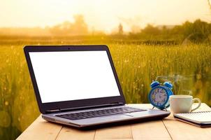 concettuale spazio di lavoro, vuoto spazio scrivania con su il computer portatile schermo e bloc notes e caffè tazza e in punto a sfocato sfondo di prati Alba estate natura sfondo. foto