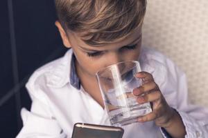 piccolo ragazzo potabile acqua e utilizzando mobile Telefono. foto