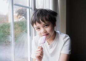 un' contento ragazzo mangiare alcuni ghiaccio crema, ritratto di un' bello giovane ragazzo seduta Il prossimo per un' finestra avendo un' rinfresco, bambino con un' sorridente viso rilassante a casa. ragazzo guardare a telecamera foto