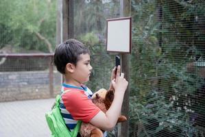 scuola ragazzo utilizzando mobile Telefono assunzione foto di animale nel il gabbia nel il azienda agricola, bambino ragazzo apprendimento di natura nel il zoo, contento ragazzo avendo divertimento con animali safari parco su caldo estate giorno