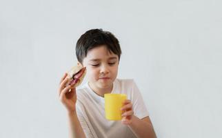 Affamato giovane ragazzo mangiare fatti in casa Bacon panini con misto la verdura, salutare ragazzo potabile succo per prima colazione a casa, bambino mordere dito Chiodi e guardare su con pensiero viso foto