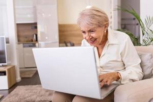 anziano signora Lavorando con il computer portatile. ritratto di bellissimo più vecchio donna Lavorando il computer portatile computer al chiuso. anziano donna utilizzando il computer portatile a casa, ridendo foto