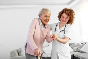 sorridente infermiera porzione anziano signora per camminare in giro il assistenza infermieristica casa. ritratto di contento femmina badante e anziano donna a piedi insieme a casa. professionale badante assunzione cura di anziano donna. foto