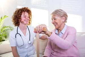 giovane badante e anziano donna ridendo insieme mentre seduta su divano. anziano donna e minore amico avendo divertimento insieme durante incontro a casa. foto