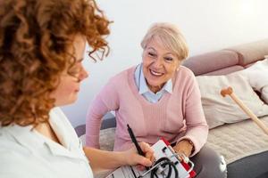 anziano donna è visitato di sua medico o Caregiver. femmina medico o infermiera parlando con anziano paziente. medicinale, età, Salute cura e casa cura concetto. anziano donna con sua badante a casa foto